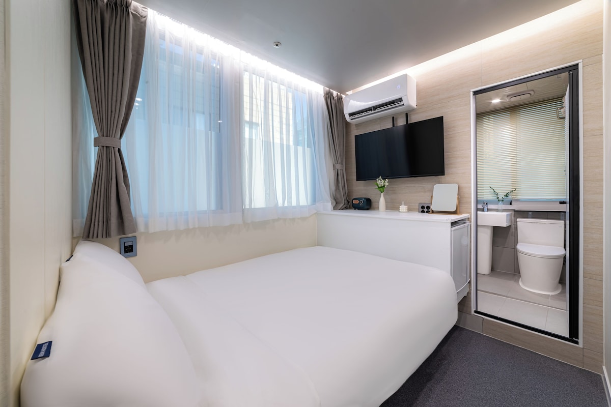 Une chambre d’hôtel comprenant un lit, une télévision, une petite table avec un vase, et une salle de bain avec des toilettes et un lavabo.