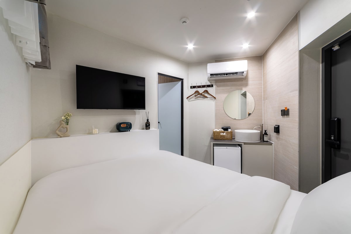 Une chambre d’hôtel moderne équipée d’un grand lit, d’un téléviseur à écran plat, d’une petite table de chevet, d’un miroir, d’un climatiseur fixé au mur et d’une porte.