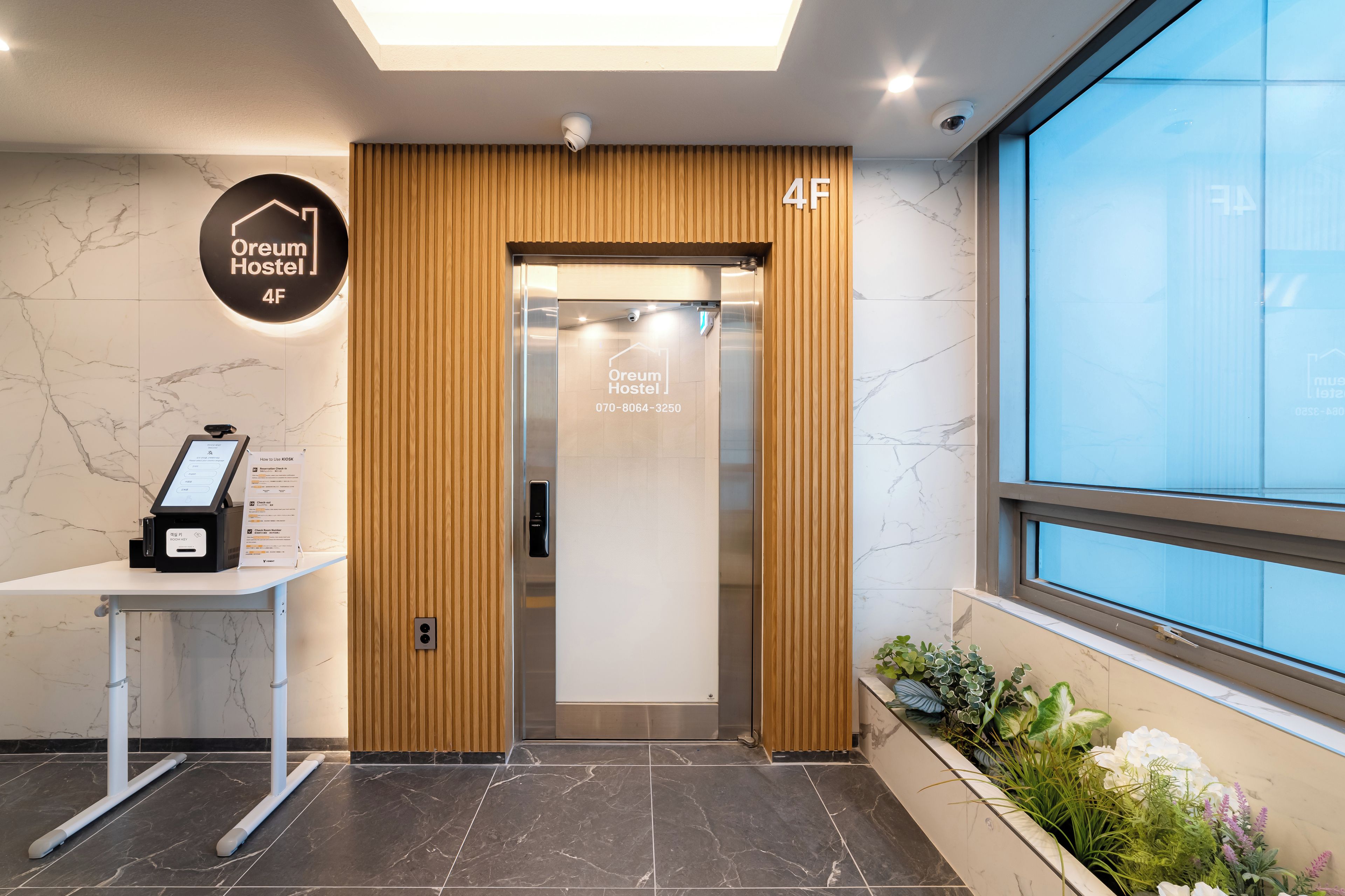 A hotel reception area with a kiosk, elevator, and a sign indicating the location as 'Oream Hostel 4F'.