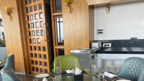 Uma área de refeições com mesa e cadeiras verdes, cozinha ao fundo e divisória de madeira com painéis de vidro.