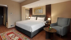 Quarto de hotel com uma cama grande, uma cadeira, uma mesinha de centro com um vaso e um quadro na parede.