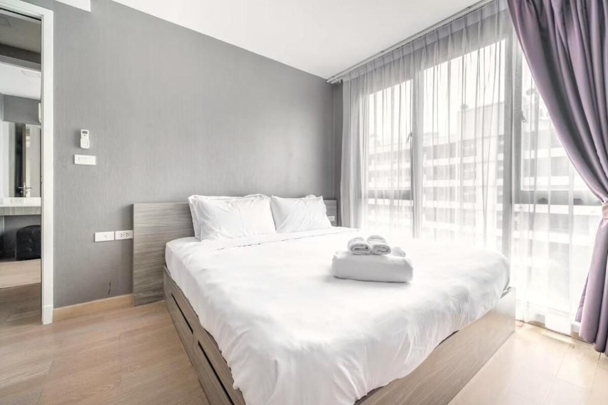 A modern hotel room with a large bed, a bedside table, a mirror, and a window with curtains.