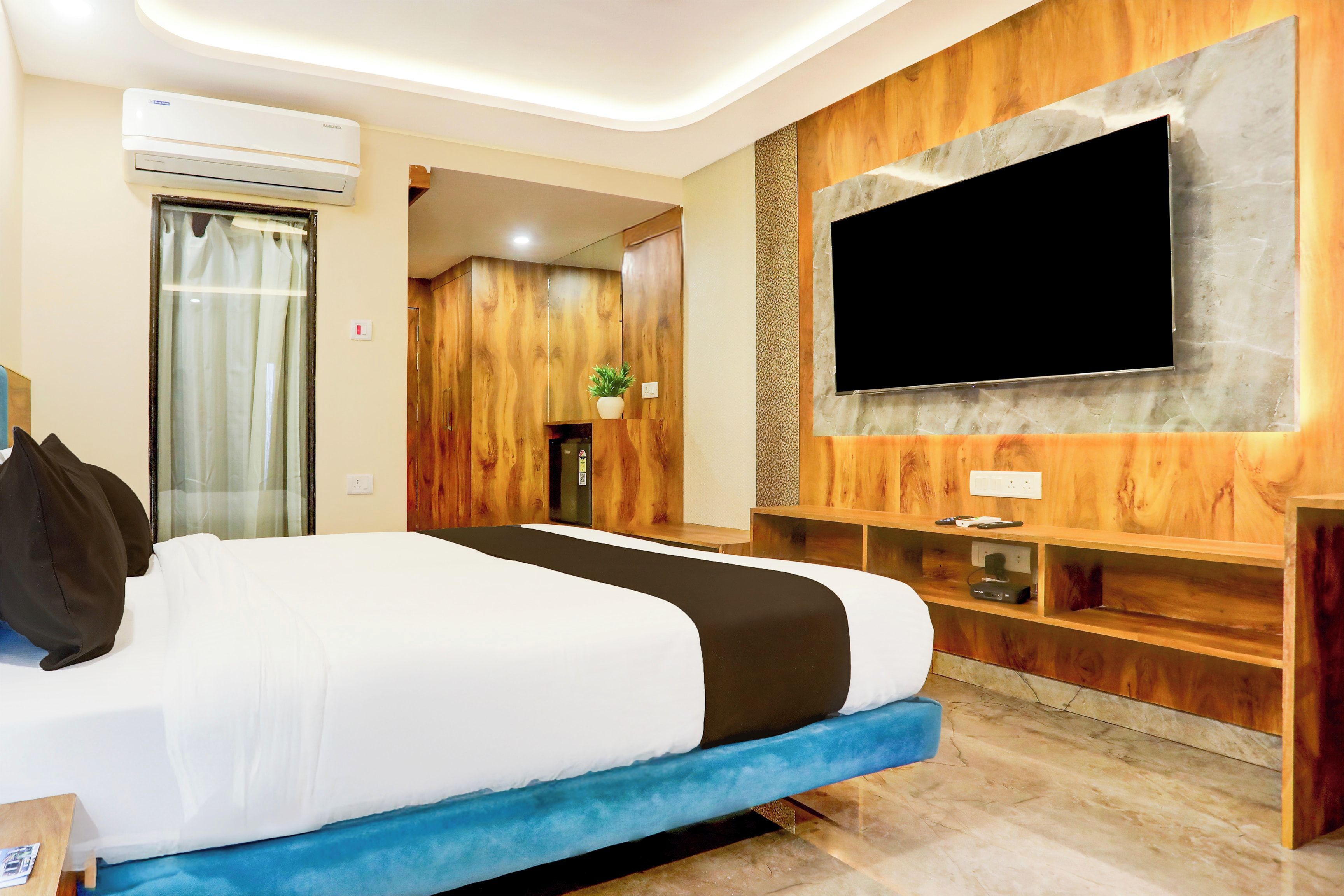 Habitación de hotel con cama, televisor de pantalla plana montado en una pared de madera, equipo de aire acondicionado y una planta pequeña en un estante.