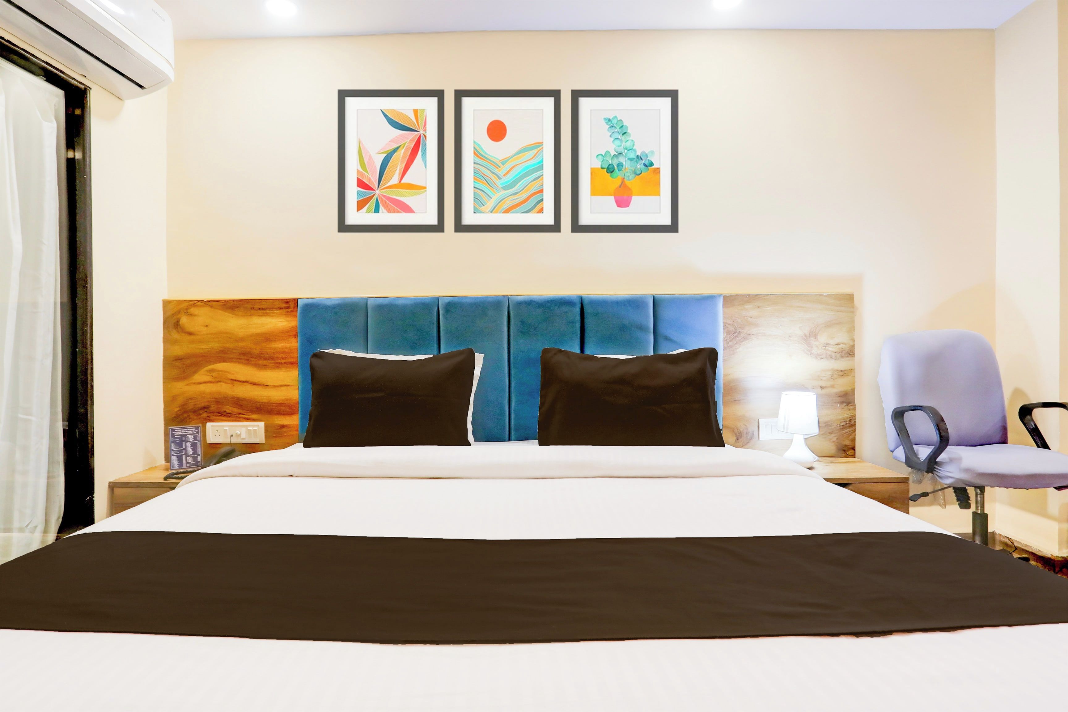 Quarto de hotel com uma cama, mesa de cabeceira, uma cadeira e três quadros na parede.