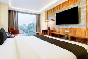 A hotel room with a large bed, a flat-screen TV mounted on the wall, a seating area with a table and chairs, and a view of the city through the window.