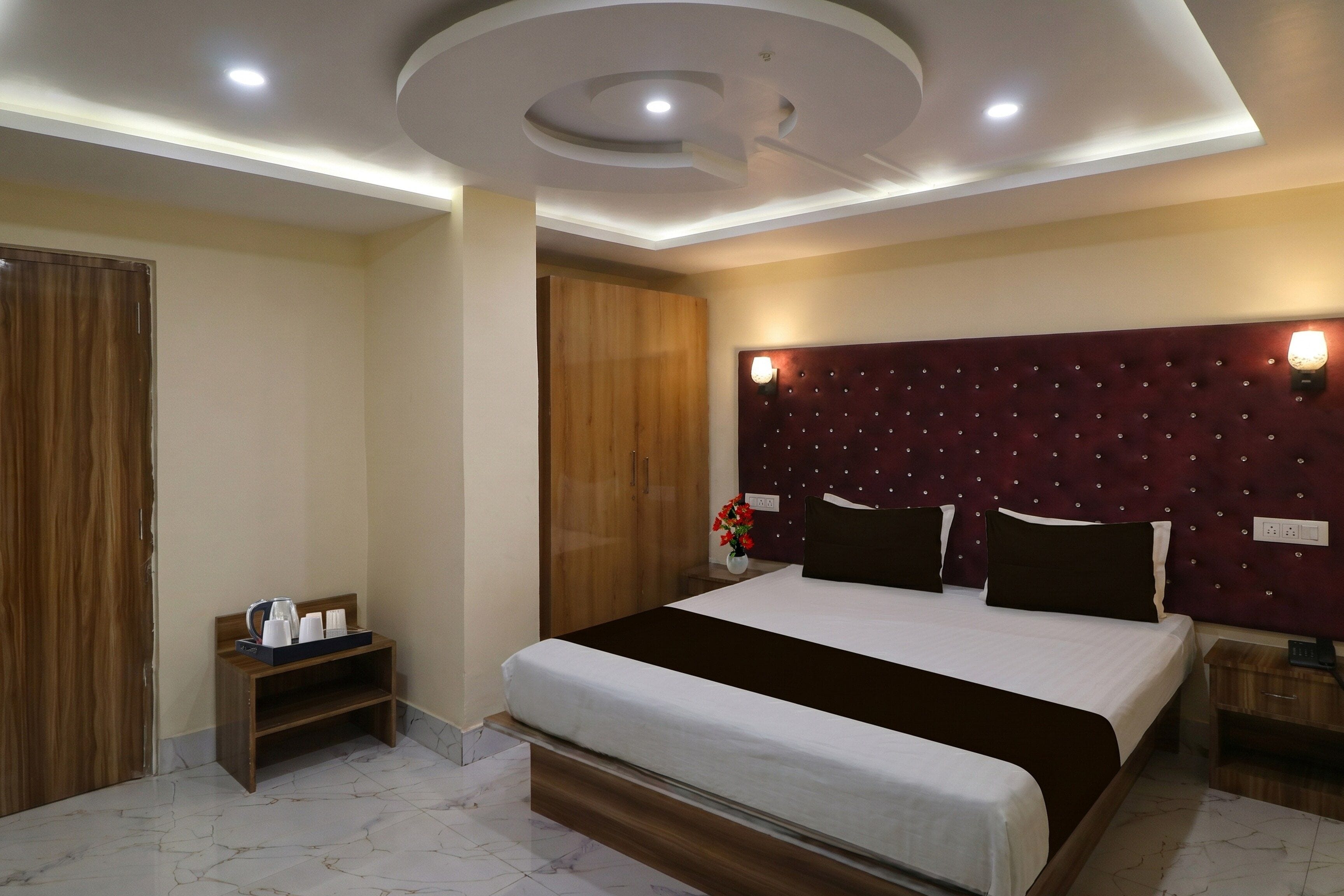 Una habitación de hotel con una cama grande, un armario de madera, una mesita de noche con cafetera y una lámpara de pared.