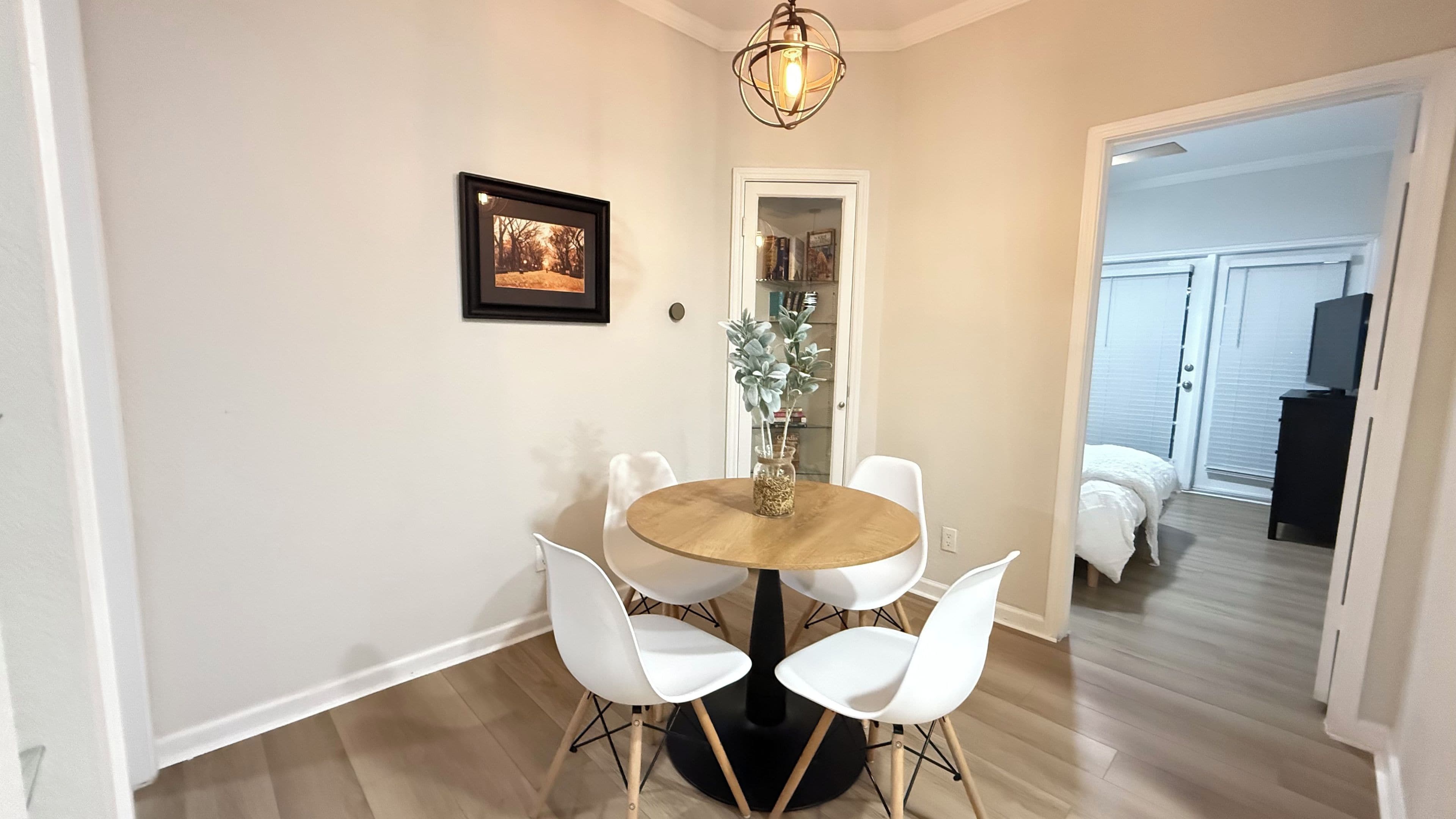 A dining area with a round table, white chairs, a vase with plants, a framed picture on the wall, and a door leading to another room.