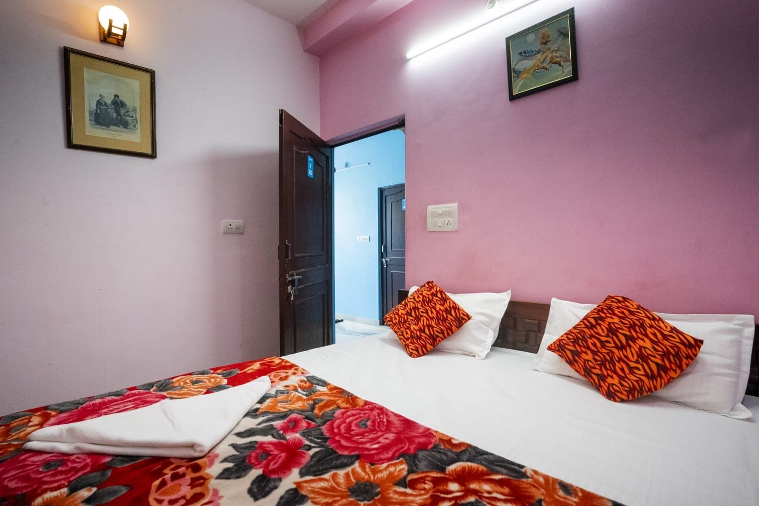 A hotel room with a bed, pillows, a wall-mounted light, and a door leading to another room.