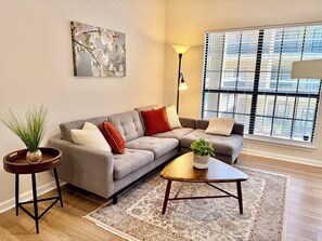 Ein modernes Wohnzimmer mit einem grauen Ecksofa, einem kleinen runden Tisch und einem raumhohen Fenster mit Jalousien.