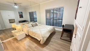 Quarto com cama, mesas de cabeceira, escrivaninha e janela com persianas.