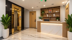 Zona de recepción de un hotel con suelo de mármol, mostrador de recepción, una planta y una estantería.