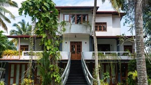 Una casa de dos pisos con una escalera central, rodeada de abundante vegetación y palmeras.