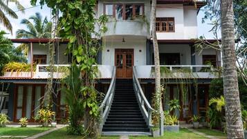 A two-story house with a central staircase, surrounded by lush greenery and palm trees.