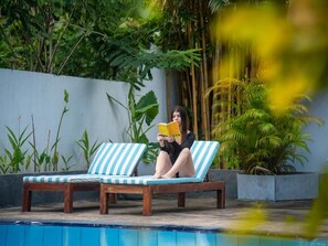 Una persona llegint un llibre en una cadira relaxant al costat d'una piscina, amb plantes tropicals al fons.