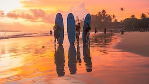 Escena de playa con surfistas sosteniendo tablas de surf, palmeras y una puesta de sol.