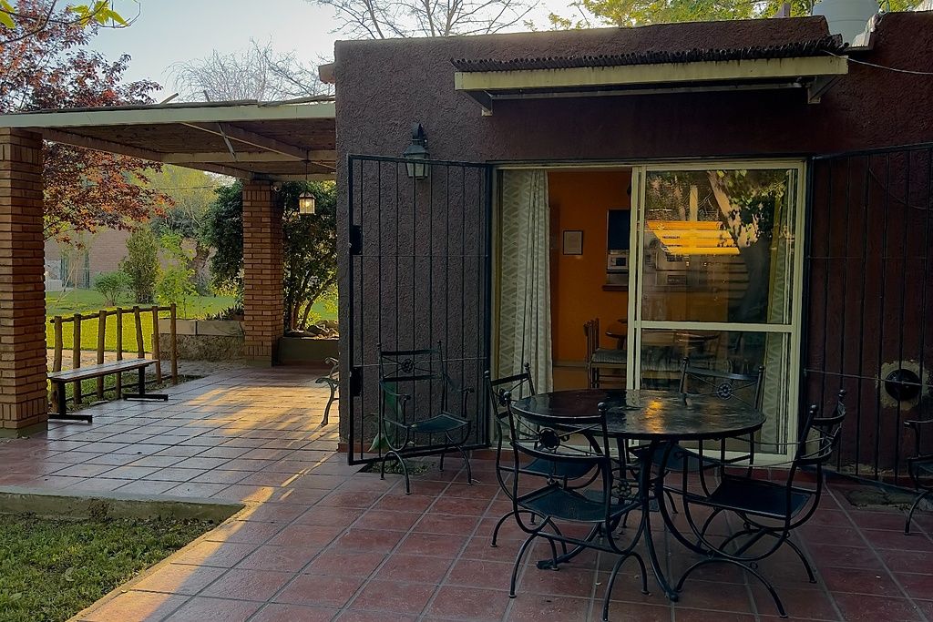 Une terrasse extérieure avec une table et des chaises, un espace salon abrité et une vue sur un bâtiment à travers une porte vitrée.