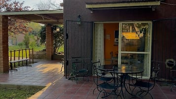 Une terrasse extérieure avec une table et des chaises, un espace salon abrité et une vue sur un bâtiment à travers une porte vitrée.