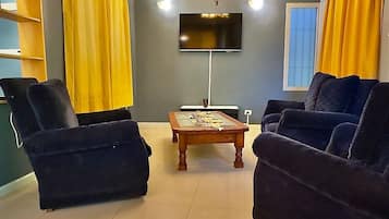 Un salon avec deux fauteuils bleu foncé, une table basse en bois, un téléviseur à écran plat fixé au mur et des rideaux jaunes.