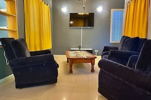 Un salon avec deux fauteuils bleu foncé, une table basse en bois, un téléviseur à écran plat fixé au mur et des rideaux jaunes.