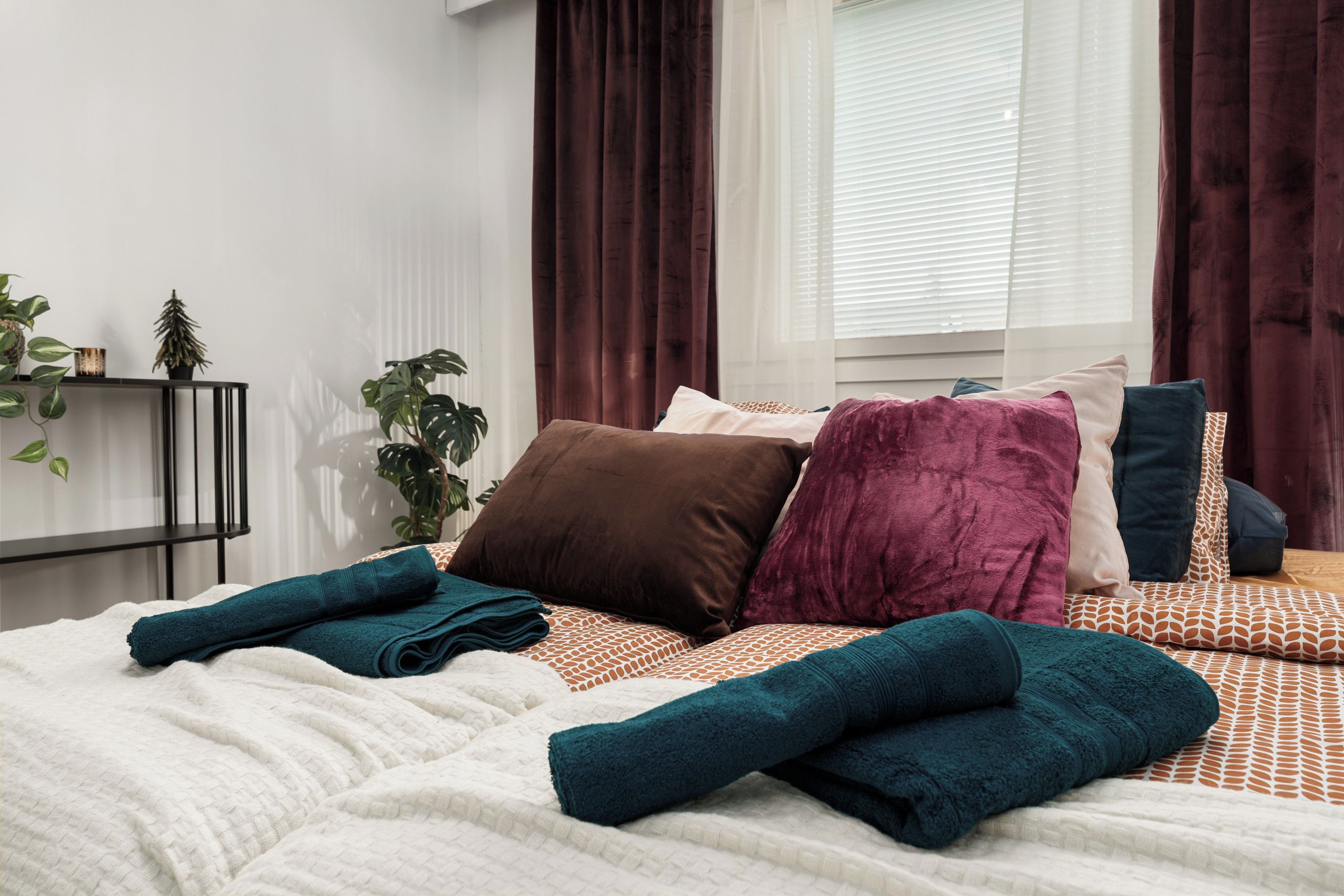 A neatly made bed with multiple pillows and folded towels, a bedside plant, and a window with curtains.