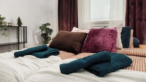 Ein ordentlich bezogenes Bett mit vielen Kissen und gefalteten Handtüchern, eine Zimmerpflanze am Nachttisch und ein Fenster mit Vorhängen.