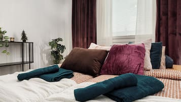 A neatly made bed with multiple pillows and folded towels, a bedside plant, and a window with curtains.