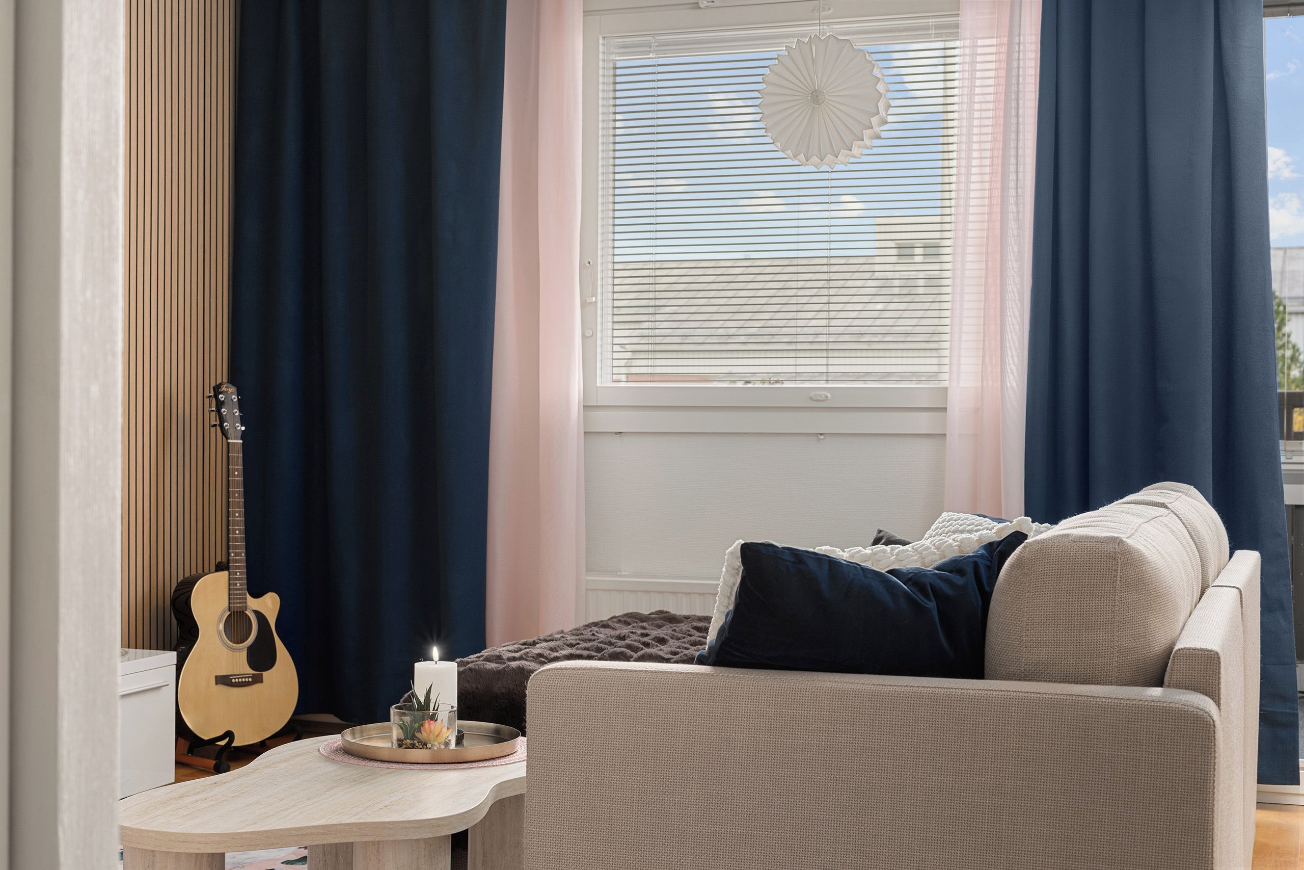 A cozy living room with a sofa, a guitar, a small table with a candle, and a window with blinds.