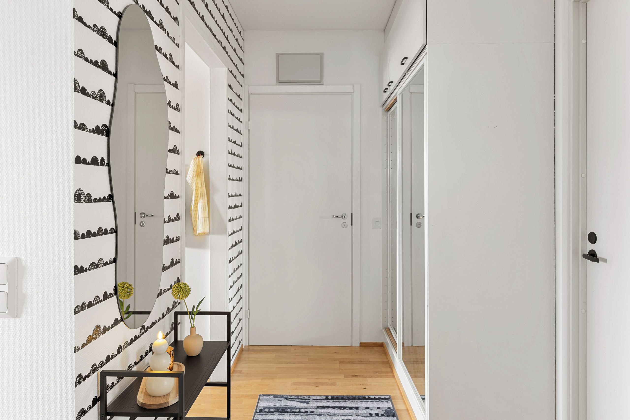 A hallway with a mirrored wall, a wooden floor, a white door, and a black-framed shelf with a vase and candle.