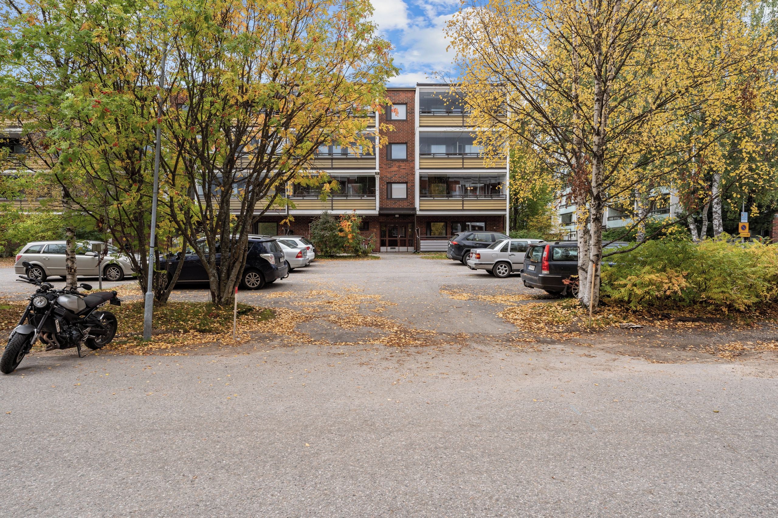 A parking area with several cars and a motorcycle, surrounded by trees with autumn leaves.