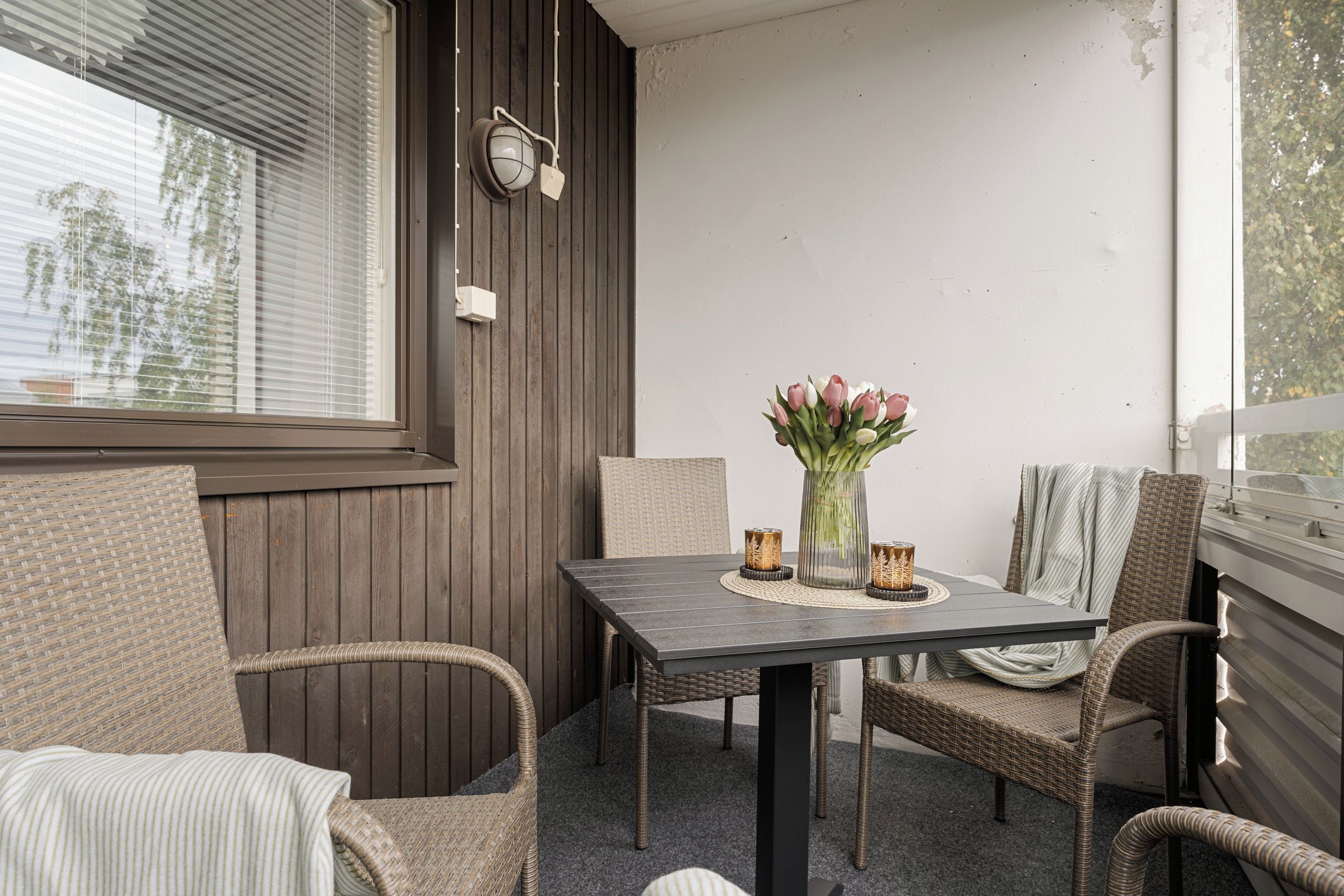 A small outdoor seating area with wicker chairs and a table, a potted plant, and a window with blinds.