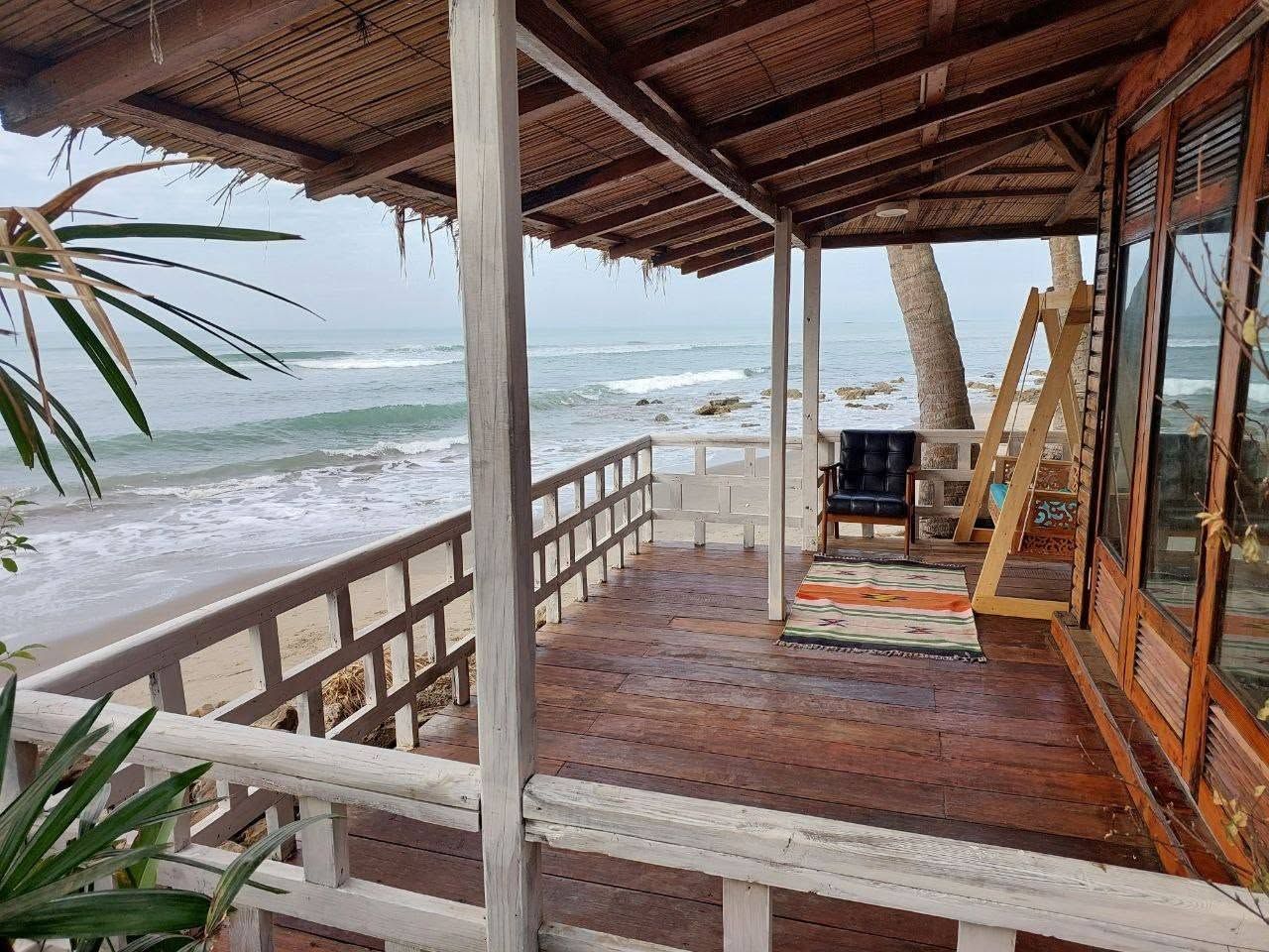 Une terrasse en bois avec une rambarde donnant sur l’océan, un espace détente avec un fauteuil, et une structure en bois dotée de fenêtres en verre.