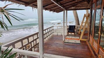 Une terrasse en bois avec une rambarde donnant sur l’océan, un espace détente avec un fauteuil, et une structure en bois dotée de fenêtres en verre.
