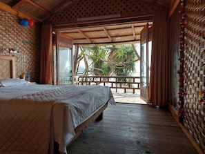 Ein Zimmer mit einem Bett, Holzboden, einem Balkon mit Blick auf Palmen und einer gewobenen Wanddekoration.