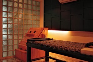 Ein Zimmer mit einem Bett, einer hölzernen Bank, einer kleinen Holzkonstruktion und einer Wand mit einer Anordnung aus Glassteinen.