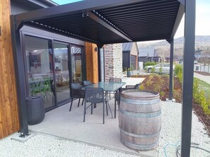 A covered outdoor seating area with a glass table and chairs, a wooden barrel, and a gravel floor. - Cromwell Central Charm. Perfect location to explore the stunning Central Otago. (Cromwell)