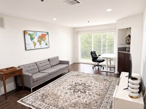 Ein modernes Wohnzimmer mit einem grauen Sofa, einem Holzbeistelltisch, einem gemusterten Teppich, einem schwarzen Bürostuhl und einem weißen Schreibtisch mit einem Globus.