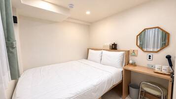 A single bed with a wooden headboard, a desk with a chair, a trash bin, a mirror, and a small table with a lamp and a mug.
