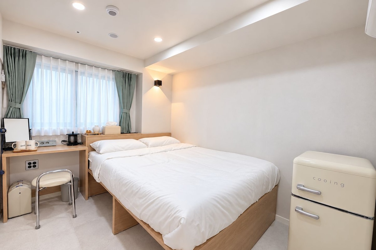 A hotel room with a bed, a desk with a chair, a small refrigerator, and a window with curtains.