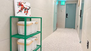 A hallway with a green metal shelf holding white baskets, a painting of red flowers on the wall, and a door labeled 'H10' with an exit sign above it. - Hub82 (Seoul)