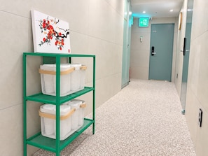 Un couloir avec une étagère métallique verte sur laquelle sont posés des paniers blancs, un tableau représentant des fleurs rouges accroché au mur, et une porte portant l’inscription « H10 », avec un panneau indiquant la sortie au-dessus.