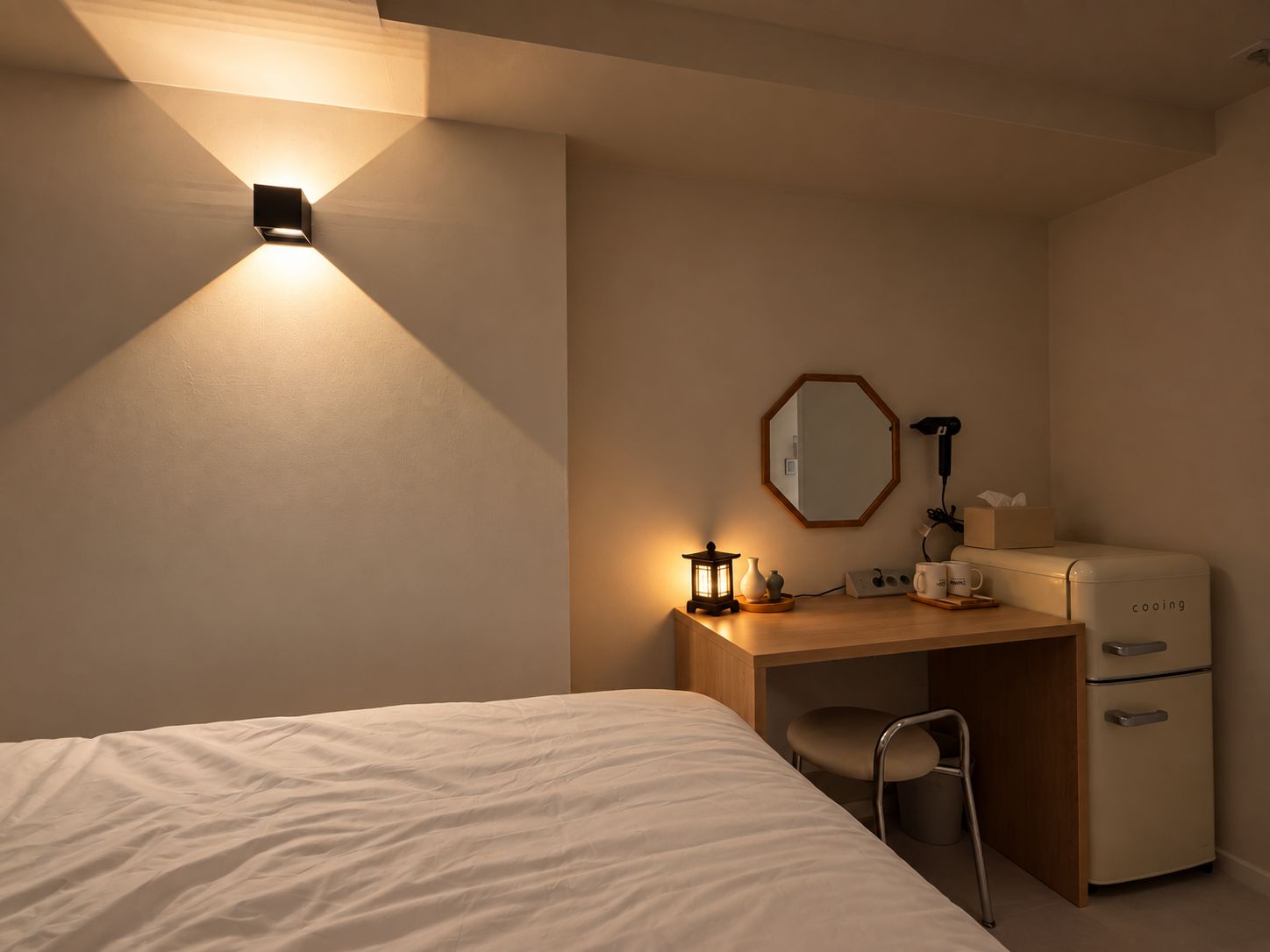 A hotel room with a bed, a desk, a chair, a small table, a lamp, a mirror, a heater, and a refrigerator.