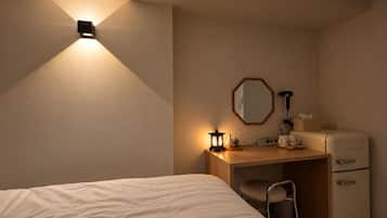 A hotel room with a bed, a desk, a chair, a small table, a lamp, a mirror, a heater, and a refrigerator.