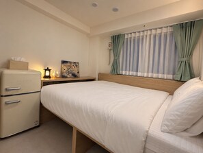 A hotel room with a bed, a small kitchenette with a fridge, a bedside table with a lamp, and a window with curtains. - Hub82 (Seoul)