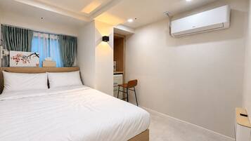 A hotel room with a bed, a desk, an air conditioning unit, and a view of a corridor.