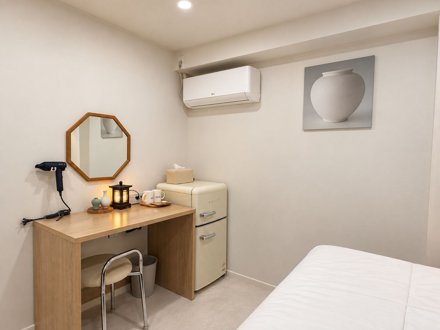A compact hotel room with a wooden desk, a small chair, a bed, an air conditioning unit, and a wall-mounted mirror.