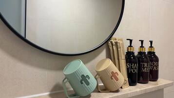 A bathroom with a round mirror, two mugs with crosses, and three bottles of shampoo.