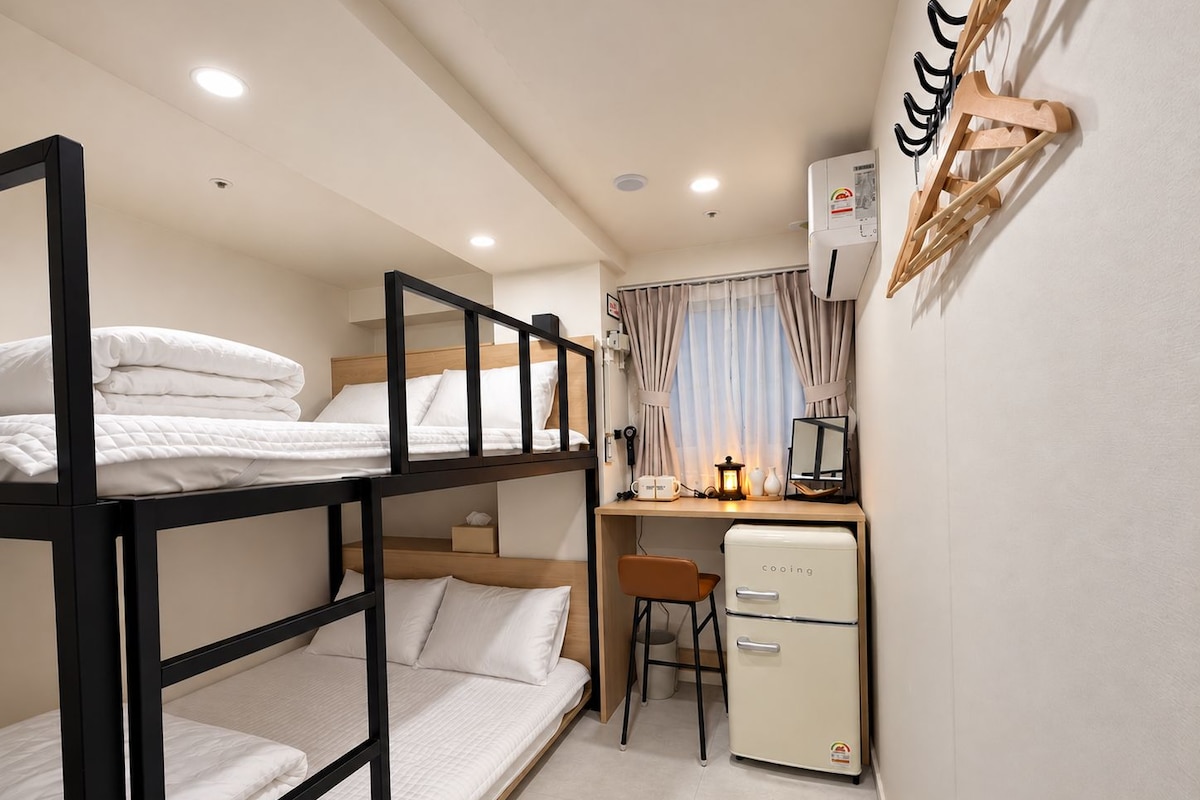 Une chambre compacte équipée de lits superposés, d’un bureau, d’une chaise, d’un petit réfrigérateur et d’un climatiseur fixé au mur.