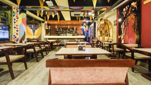 Een restaurantinterieur met houten tafels en banken, een bar en kleurrijke wanddecoraties.