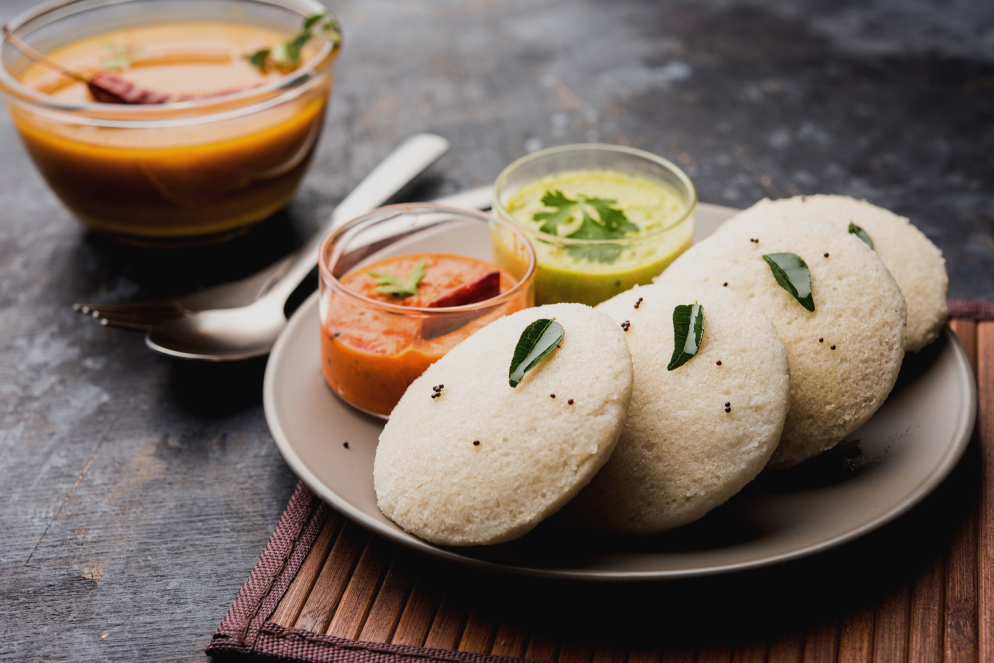Un plato con idlis y tres chutneys diferentes, un tazón de sopa y una cuchara sobre una superficie de madera.
