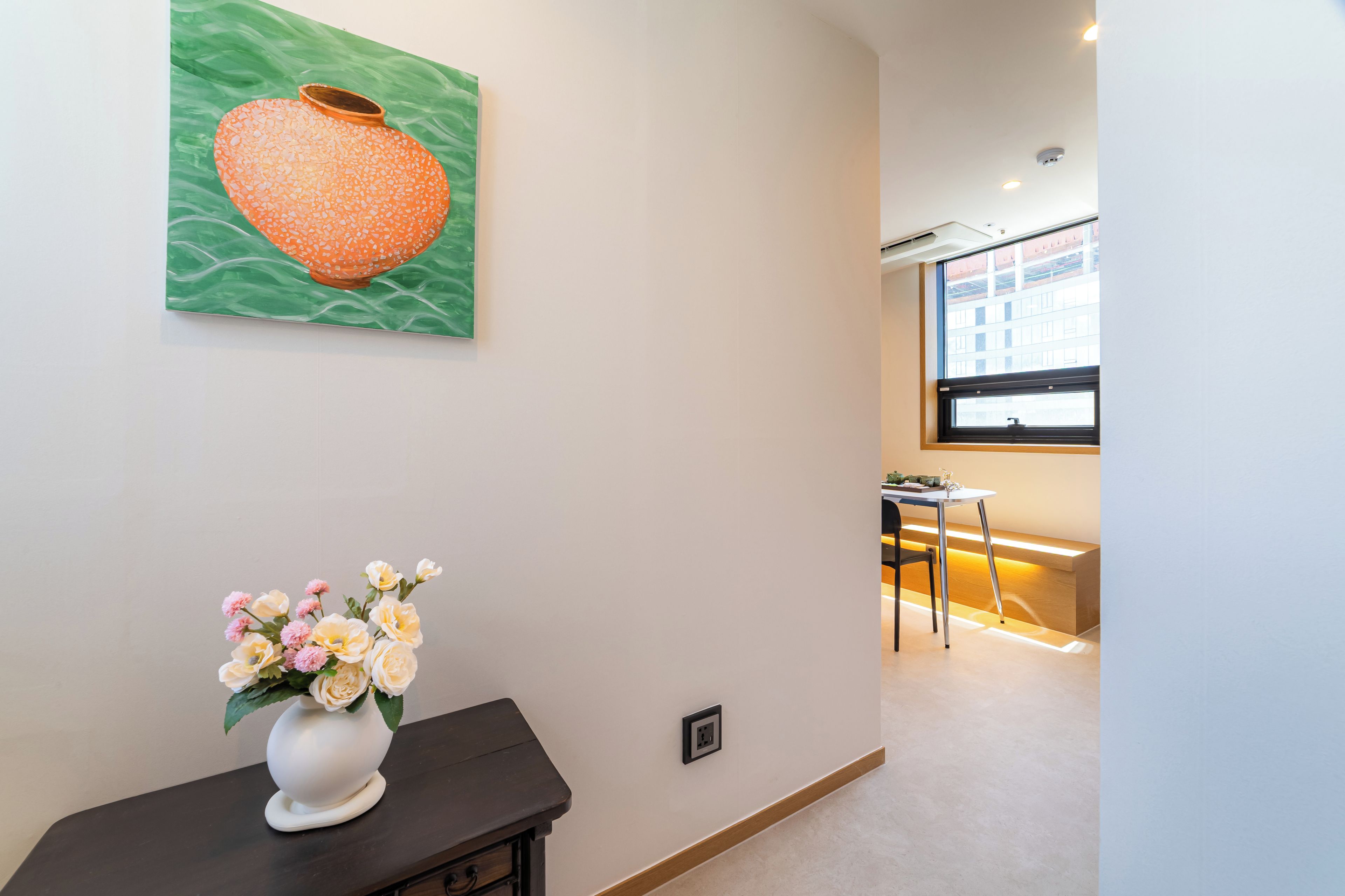 Un interior moderno con un florero sobre una mesa oscura, un cuadro de una maceta de cerámica en la pared, y una vista parcial de un comedor con una ventana.